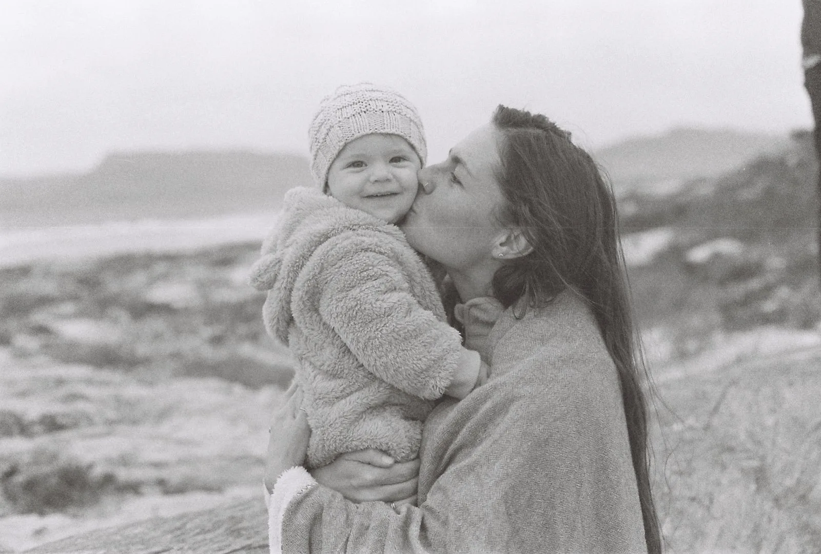 A mother kissing her bundled-up baby on a windswept beach